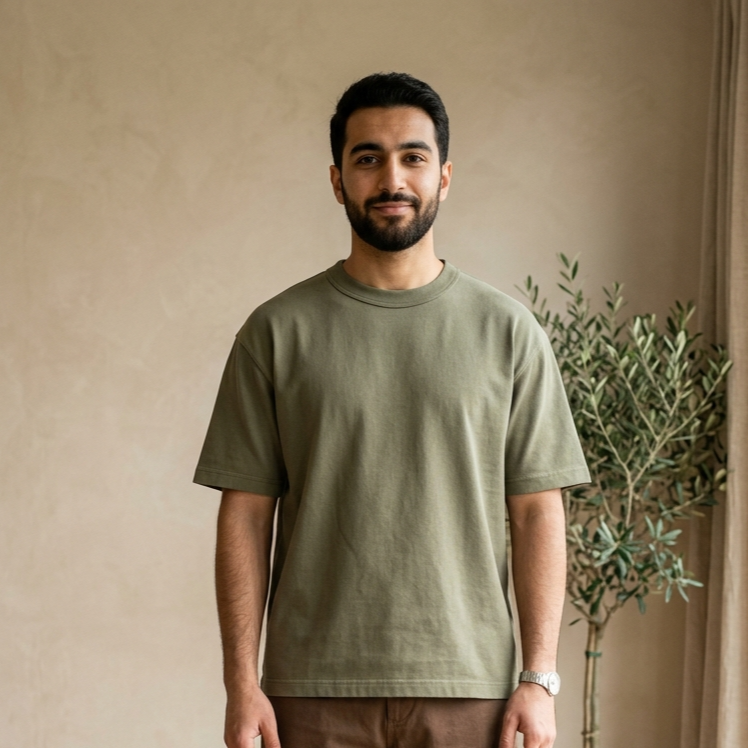Man wearing a green t-shirt and brown pants standing in a room with a plant and wooden floor.